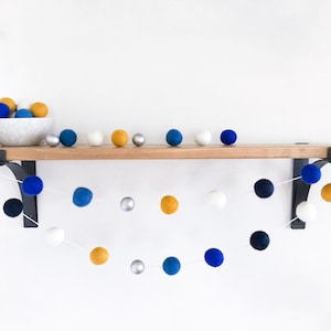 May include: A garland of felt balls in blue, white, gold, and silver colors hangs from a wooden shelf. The garland is made of felt balls strung together with white thread.