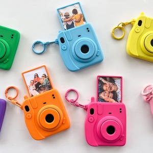 May include: A collection of colorful, miniature camera-shaped keychains in green, blue, yellow, orange, purple, pink. Each keychain has a circular lens and a small photo displayed. The keychains are arranged on a white surface.