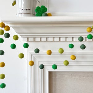 May include: A green and yellow felt ball garland hangs over a white fireplace mantel. The garland is made of small, round felt balls in shades of green and yellow.