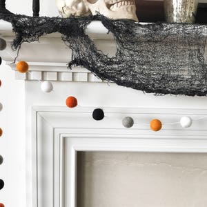 May include: A Halloween garland with black, white, orange, and gray felt balls strung on a white string. The garland is hanging on a white fireplace mantel with a black spiderweb draped over it.