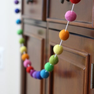 May include: A rainbow felt ball garland hanging on a wooden cabinet door. The garland is made of colorful felt balls strung together with white thread.