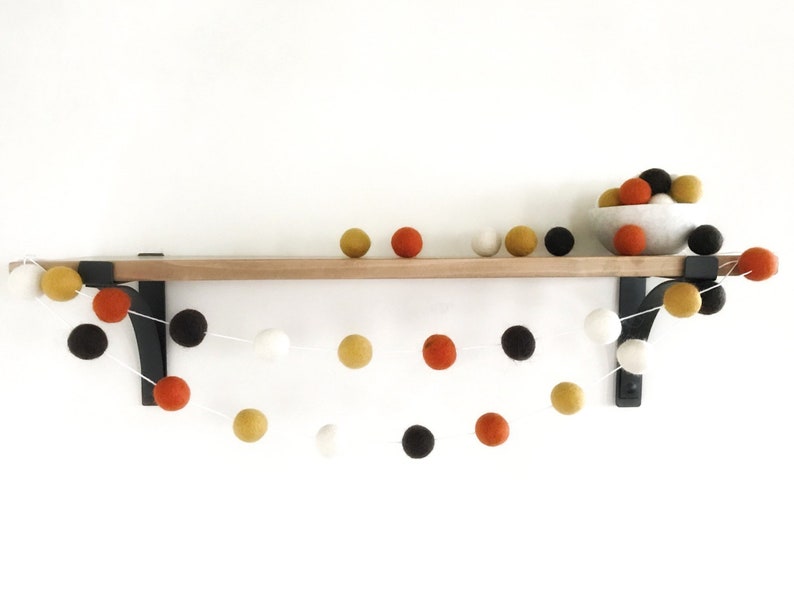 May include: A garland of felt balls in orange, white, black, and gold colors hangs from a wooden shelf with black metal brackets.