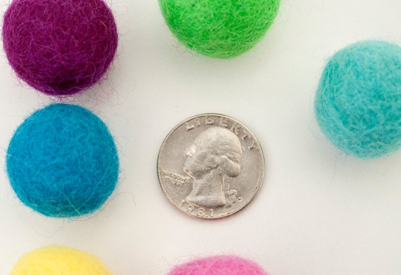 May include: A collection of colorful felt balls in shades of purple, green, blue, yellow, and pink, arranged around a silver US quarter with the year 1981 visible.