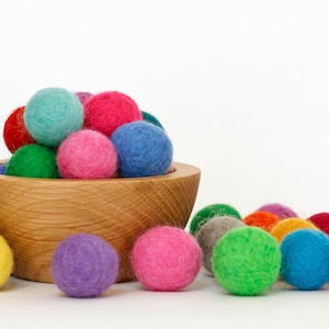 May include: A wooden bowl filled with colorful felt balls. The bowl is sitting on a white surface. There are more felt balls scattered around the bowl.