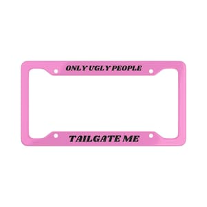 Puede incluir: Un marco rosa para matrícula con las frases "ONLY UGLY PEOPLE" y "TAILGATE ME" en texto negro. El marco tiene forma rectangular con esquinas redondeadas y orificios de montaje.