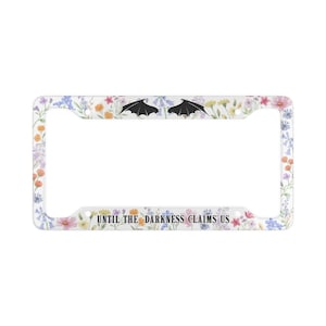 Può includere: Una cornice bianca per targa con un design floreale e la scritta "UNTIL THE DARKNESS CLAIMS US". La cornice presenta ali di pipistrello nere nella parte superiore e fiori colorati sui bordi.
