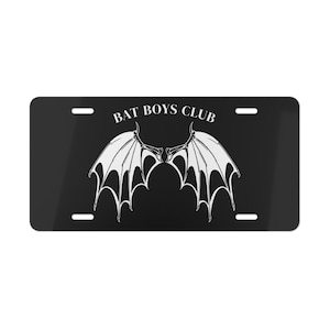 Op de afbeelding: Zwarte nummerplaat met de tekst "BAT BOYS CLUB" boven een witte afbeelding van vleermuisvleugels. De vleugels zijn symmetrisch en gedetailleerd, met een donkere achtergrond en witte accenten.