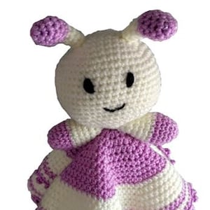Pode incluir: Um animal de pelúcia branco de crochê com orelhas roxas e um cobertor listrado roxo e branco. O animal tem um rosto sorridente com olhos pretos.