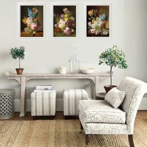May include: Three framed floral still life paintings with white frames and a white background. The paintings are hanging on a white wall above a wooden console table with two striped ottomans and a chair with a floral patterned upholstery.