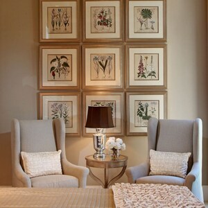 May include: Nine framed botanical prints with gold-colored frames are displayed on a beige wall. Two gray upholstered armchairs with white pillows flank a small table with a lamp and flowers. A bed with a beige cover and a pink throw is in the foreground.