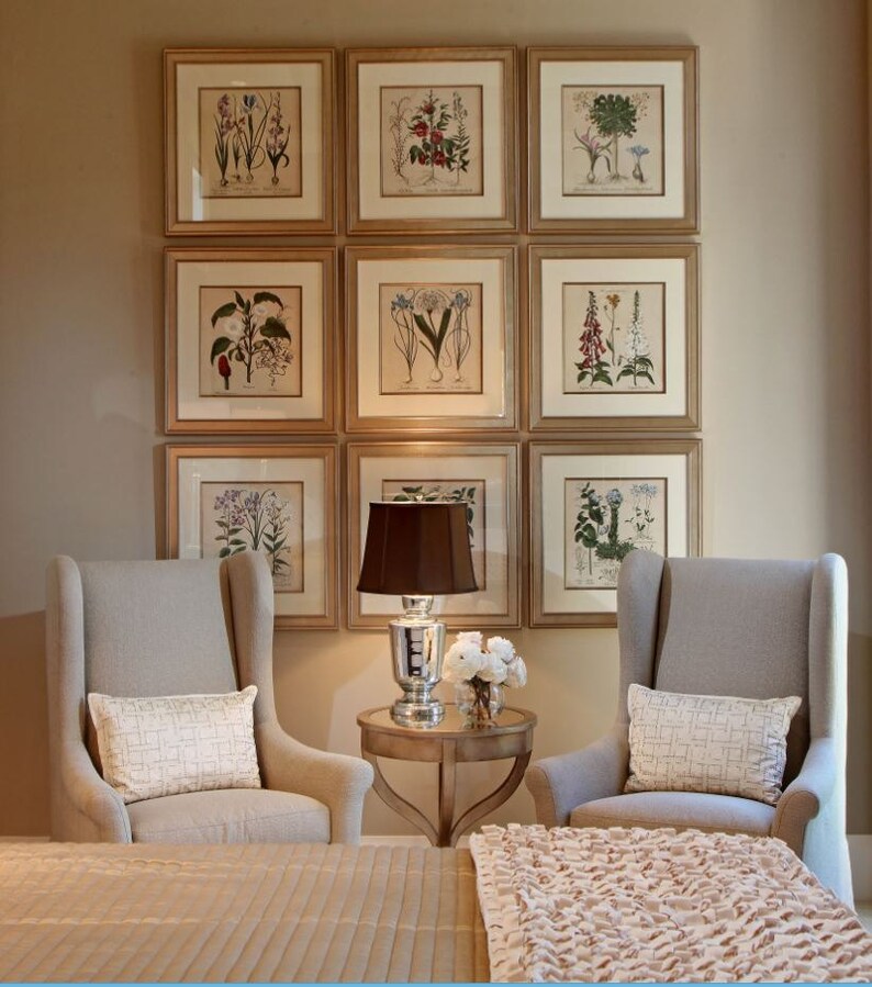 May include: Nine framed botanical prints with gold-colored frames are displayed on a beige wall. Two gray upholstered armchairs with white pillows flank a small table with a lamp and flowers. A bed with a beige cover and a pink throw is in the foreground.
