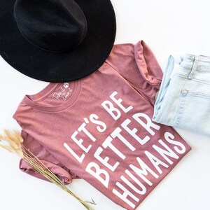May include: A mauve t-shirt with the text "Let's Be Better Humans" printed in white. The shirt is folded and laid on a white surface with a black hat and blue jeans.
