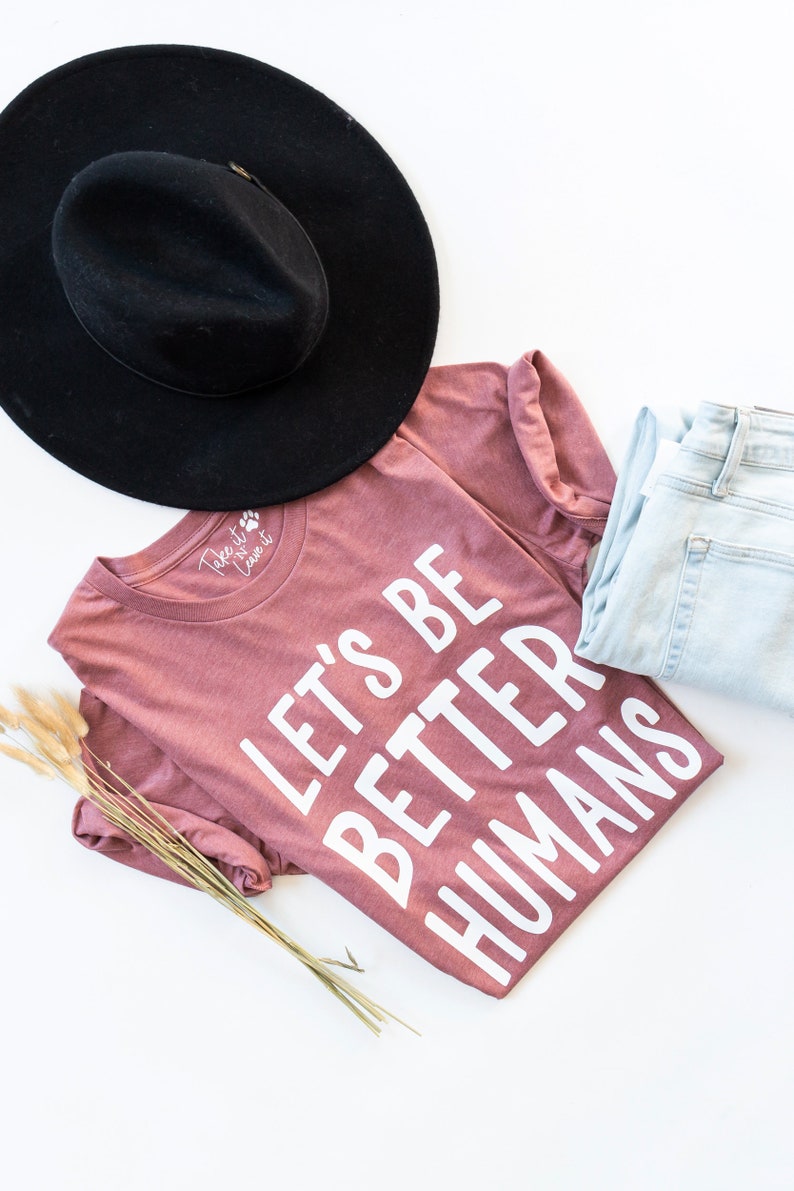 May include: A mauve t-shirt with the text "Let's Be Better Humans" printed in white. The shirt is folded and laid on a white surface with a black hat and blue jeans.