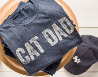May include: A dark blue t-shirt with the text "CAT DAD" printed in white on the front. The shirt is folded and sitting on a wooden surface.