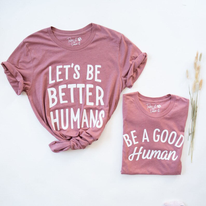 May include: Two pink t-shirts with white text. The larger shirt says "Let's Be Better Humans". The smaller shirt says "Be A Good Human".