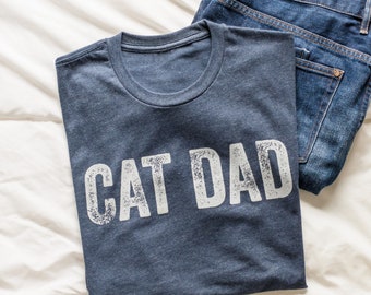May include: A dark blue t-shirt with the text "CAT DAD" printed in white on the front. The shirt is folded and laid on a white surface.