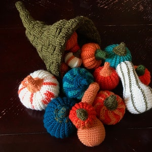 May include: A collection of crocheted pumpkins and gourds in various colours, including orange, white, green, and blue, spilling out of a green crocheted cornucopia.