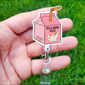 May include: Pink and white cartoon juice box with a goose and the text "Silly Goose Juice" and "Contains 100% Silly". The juice box is attached to a white retractable badge holder with a clear plastic strap and a sparkly gem.