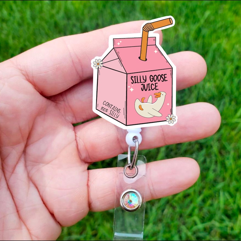May include: Pink and white cartoon juice box with a goose and the text "Silly Goose Juice" and "Contains 100% Silly". The juice box is attached to a white retractable badge holder with a clear plastic strap and a sparkly gem.