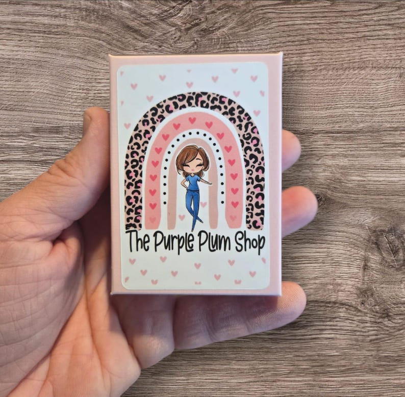 May include: A pink box with a white sticker featuring a rainbow with a leopard print design. A cartoon nurse stands in front of the rainbow. The text "The Purple Plum Shop" is printed below the rainbow.