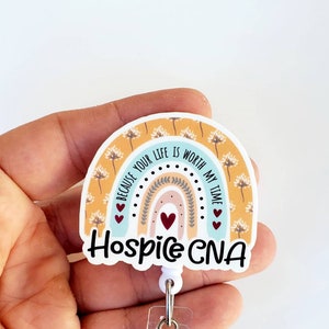 May include: A white rainbow-shaped badge holder with the text "Because your life is worth my time" and "Hospice CNA" in black lettering. The rainbow has a yellow background with white dandelion patterns and a blue, pink, and peach gradient. There are three red hearts on the badge holder.