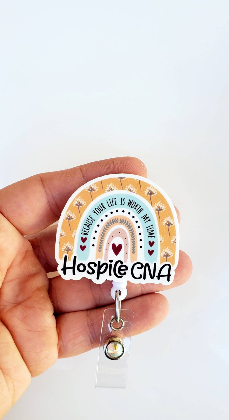 May include: A white rainbow-shaped badge holder with the text "Because your life is worth my time" and "Hospice CNA" in black lettering. The rainbow has a yellow background with white dandelion patterns and a blue, pink, and peach gradient. There are three red hearts on the badge holder.