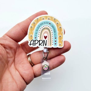 May include: A rainbow-themed badge holder with the text "LET ALL YOU DO BE DONE IN LOVE 1 CORINTHIANS 16:14" and the word "APRN" in the centre. The badge holder has a clear plastic retractable reel with a rainbow-coloured rhinestone.