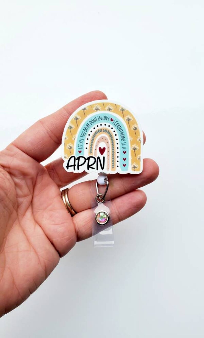May include: A rainbow-themed badge holder with the text "LET ALL YOU DO BE DONE IN LOVE 1 CORINTHIANS 16:14" and the word "APRN" in the centre. The badge holder has a clear plastic retractable reel with a rainbow-coloured rhinestone.