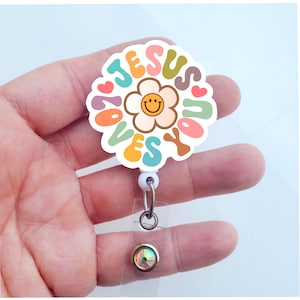 May include: A white round badge holder with a colorful flower design and the text "Jesus Loves You". The flower has a yellow center with a smiley face. The badge holder is attached to a clear plastic retractable badge reel with a silver crystal.