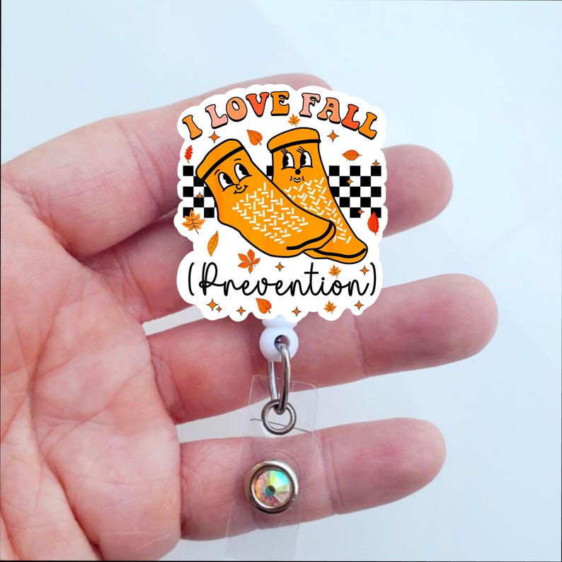 May include: A white retractable badge holder with a round orange and white sticker featuring a cartoon image of two orange socks with faces and the text "I LOVE FALL (Prevention)".