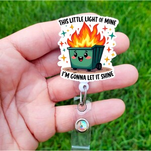 Op de afbeelding: Een witte intrekbare badgehouder met een groene cartoon vuilnisbak met vlammen erop. De vuilnisbak heeft een glimlachend gezicht en de tekst "This little light of mine I'm gonna let it shine".