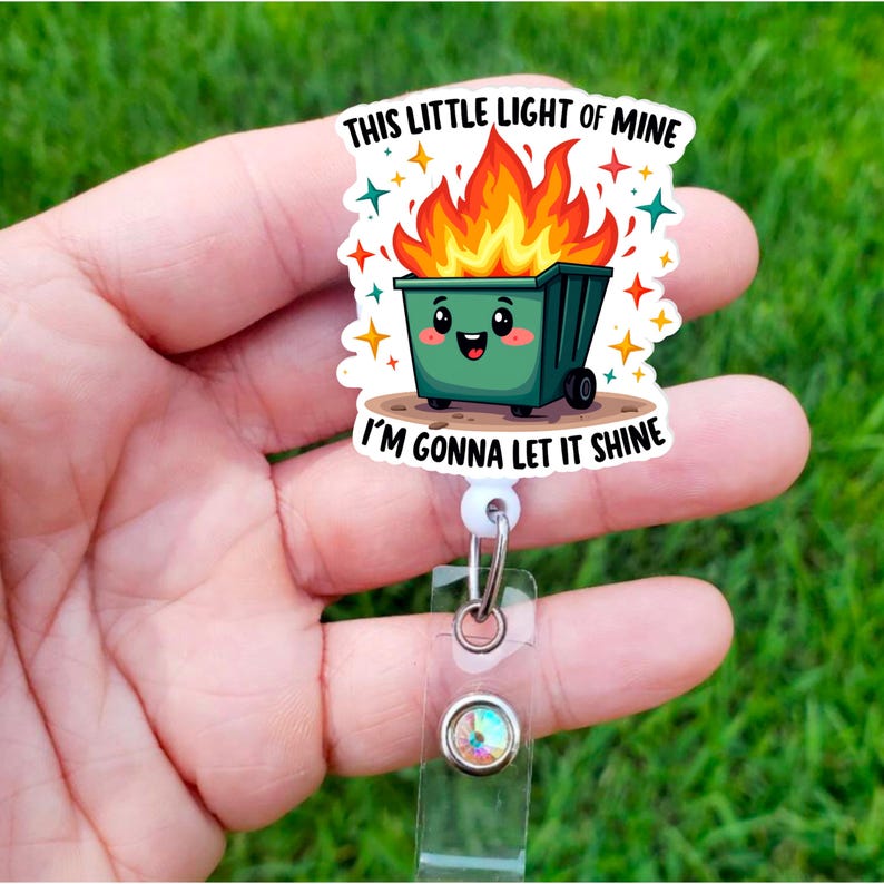 Op de afbeelding: Een witte intrekbare badgehouder met een groene cartoon vuilnisbak met vlammen erop. De vuilnisbak heeft een glimlachend gezicht en de tekst "This little light of mine I'm gonna let it shine".