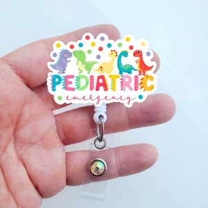 May include: A white plastic badge holder with a colorful dinosaur design and the text "PEDIATRIC emergency". The badge holder has a clear plastic retractable reel with a crystal-like button.