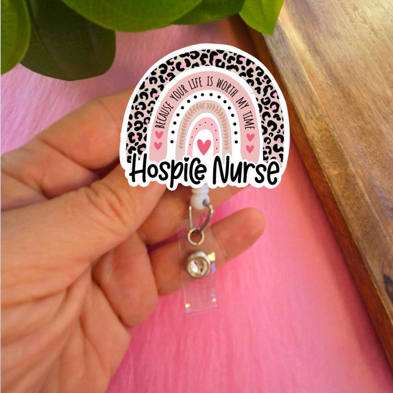 May include: A pink and black leopard print rainbow with the text "Because your life is worth my time" and "Hospice Nurse". The rainbow is attached to a clear plastic retractable badge holder with a silver rhinestone.