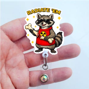 May include: A badge reel featuring a cartoon raccoon wearing sunglasses and a red shirt with a radiation symbol. The raccoon holds a bone and the text "RADIATE 'EM" is above it. The reel has a clear plastic strap and a decorative gem.
