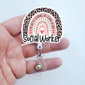 May include: A pink and black leopard print rainbow with the text "The art of listening, the science of hope" and "Social Worker" on a white background. The rainbow is attached to a white retractable badge holder with a clear plastic badge holder and a sparkly iridescent button.
