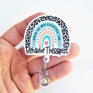 May include: A white and blue rainbow with a leopard print border and the text "Behavior Goes Where Reinforcement Flows" and "Behavior Therapist". The rainbow is attached to a clear retractable badge holder with a silver clip and a clear crystal.