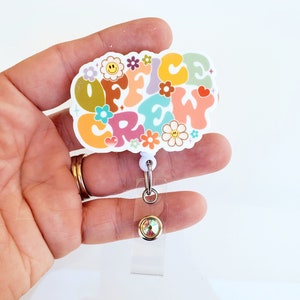 May include: A white plastic badge holder with a colorful retro design featuring the text "Office Crew" surrounded by flowers and hearts. The badge holder has a clear plastic strap and a silver retractable reel with a clear crystal.