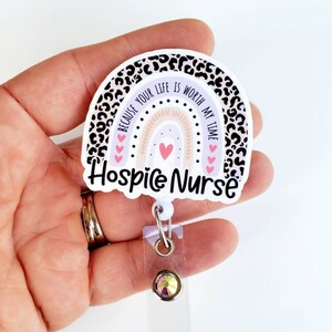 May include: A white and purple rainbow with a leopard print border. The rainbow has a pink heart in the center and the text "Because your life is worth my time" around it. The text "Hospice Nurse" is below the rainbow. The rainbow is attached to a clear plastic retractable badge holder with a sparkly rhinestone.