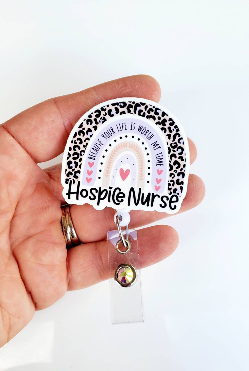 May include: A white and purple rainbow with a leopard print border. The rainbow has a pink heart in the center and the text "Because your life is worth my time" around it. The text "Hospice Nurse" is below the rainbow. The rainbow is attached to a clear plastic retractable badge holder with a sparkly rhinestone.