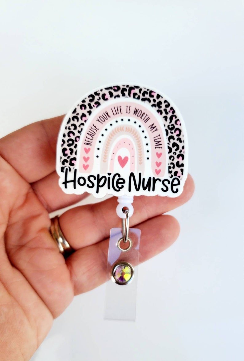 May include: White badge reel with a pink and black leopard print rainbow design. The rainbow has a pink and white background with a gold outline. The text on the badge reel reads "Because your life is worth my time." and "Hospice Nurse".