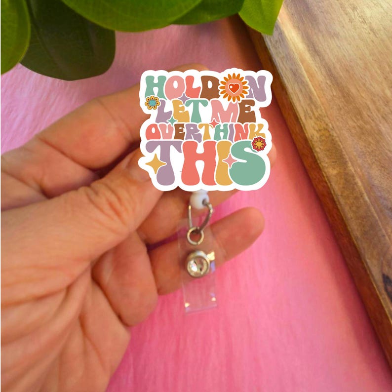 May include: A colorful, retro-style badge holder with the text "Hold on let me overthink this". The badge holder is attached to a clear plastic retractable badge reel with a silver metal clip and a clear rhinestone.