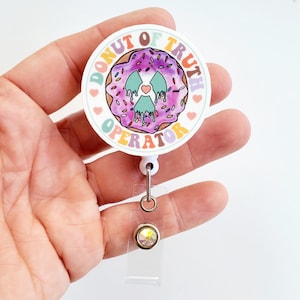 May include: A white round badge holder with a colorful donut graphic and the text "Donut of Truth Operator". The donut is purple with green icing and sprinkles. The badge holder has a clear plastic retractable reel.