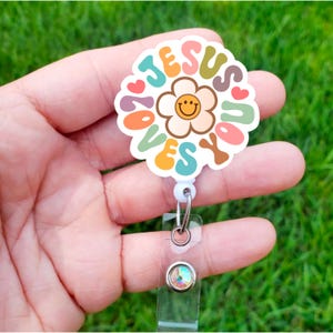 May include: A white plastic badge reel with a colorful flower design and the text "Jesus Loves You". The flower has a yellow smiley face in the center.