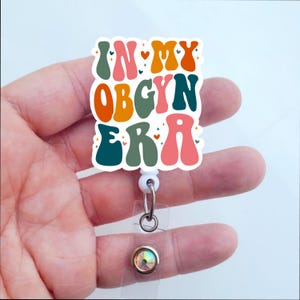 May include: A white badge holder with a colorful retro design that says "In My Obgyn Era". The badge holder has a clear plastic strap and a sparkly rhinestone.