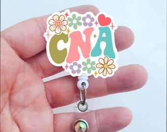 May include: A white retractable badge holder with a colorful floral design featuring the word "CNA" and a small red heart. The badge holder has a clear plastic cover and a sparkly iridescent gem.