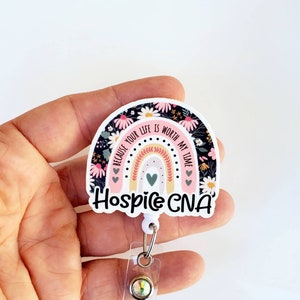 May include: A white and pink rainbow with black floral print and the text "Because your life is worth my time" and "Hospice CNA". The rainbow is attached to a clear retractable badge holder with a sparkly gem.