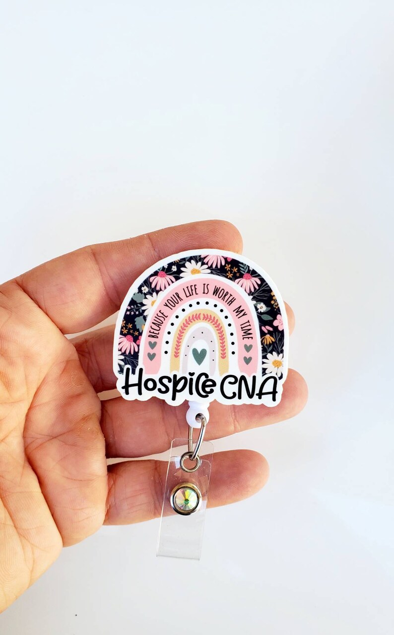 May include: A white and pink rainbow with black floral print and the text "Because your life is worth my time" and "Hospice CNA". The rainbow is attached to a clear retractable badge holder with a sparkly gem.
