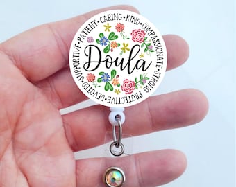 Könnte beinhalten: Runder weißer Badgehalter mit floralem Design und dem Wort "Doula" in schwarzer Schrift. Der Badgehalter hat eine weiße einziehbare Schnur und einen transparenten Plastikclip mit einem kristallartigen Anhänger.