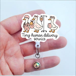 May include: A white badge reel with a cartoon design featuring three geese in medical attire. The text "Tiny human delivery service" is printed below the geese. The reel has a clear plastic strap and a decorative, iridescent button.